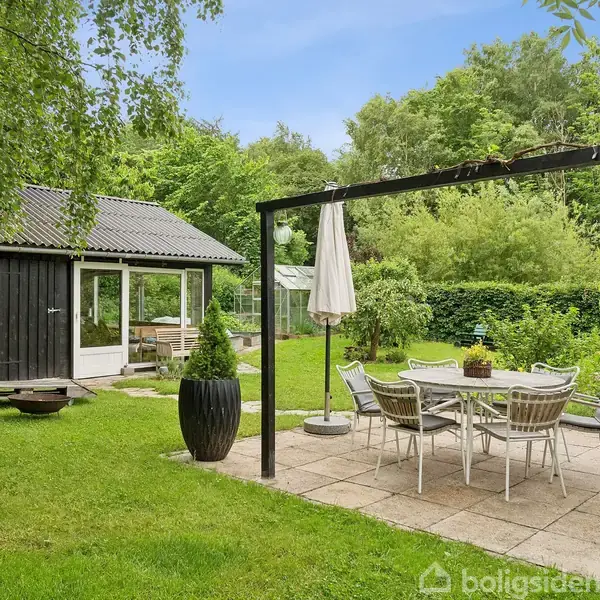 Et sort-hvidt hus med skråt tag står i en grøn have. Der er en stentrappe langs siden, en grusbelagt parkeringsplads foran og møbler på terrassen.
