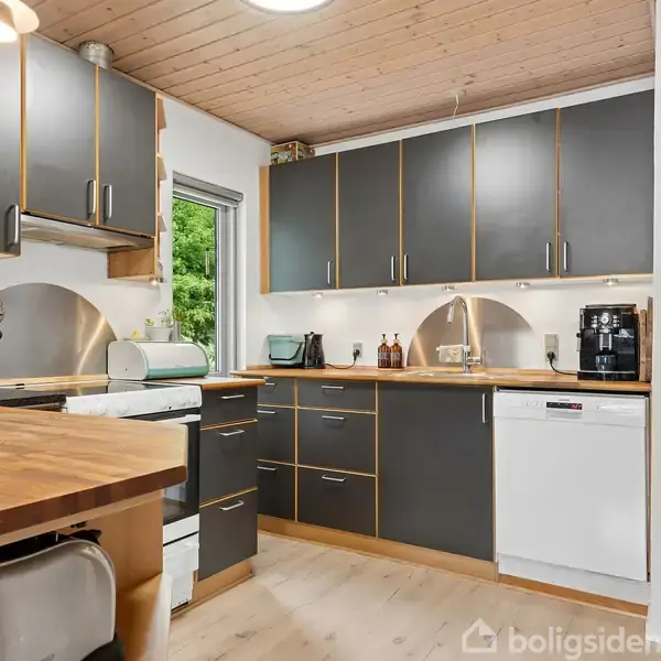 Sort-hvidt hus står i en grusbelagt gårdsplads. Huset har store vinduer, en garageport og er omgivet af frodige grønne buske og træer. I baggrunden ses endnu en bygning.