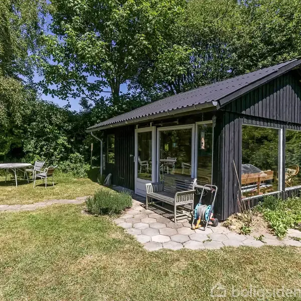 Et sort træhus står stille i en frodig have med græs, buske og træer. Foran huset er en havestol og en brolagt sti, der fører til et bord med stole.