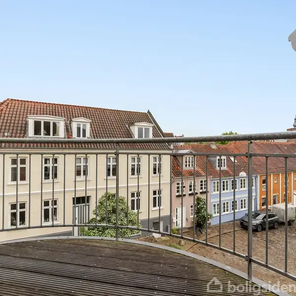 En balkon med gelænder er synlig i forgrunden; den åbner ud til en udsigt over en brostensbelagt gade med farverige, historiske bygninger i en by.