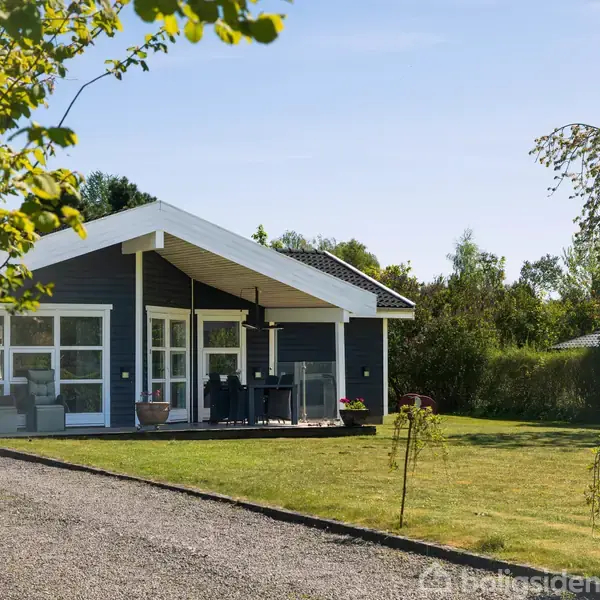 Et moderne sommerhus står på en velplejet græsplæne omgivet af træer. Huset har store vinduer og en åben terrasse med havemøbler, som åbner ud mod en grussti i forhaven.