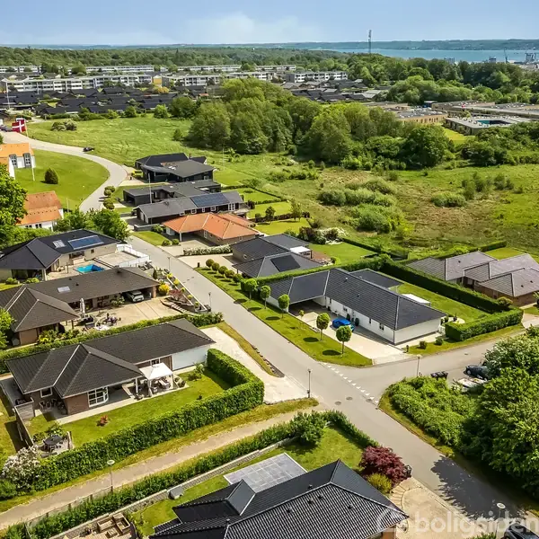 En række moderne enfamiliehuse ligger langs en snoet vej omkranset af velplejede haver. Omgivelserne består af grønne områder og i det fjerne ses en bro og en fjord.