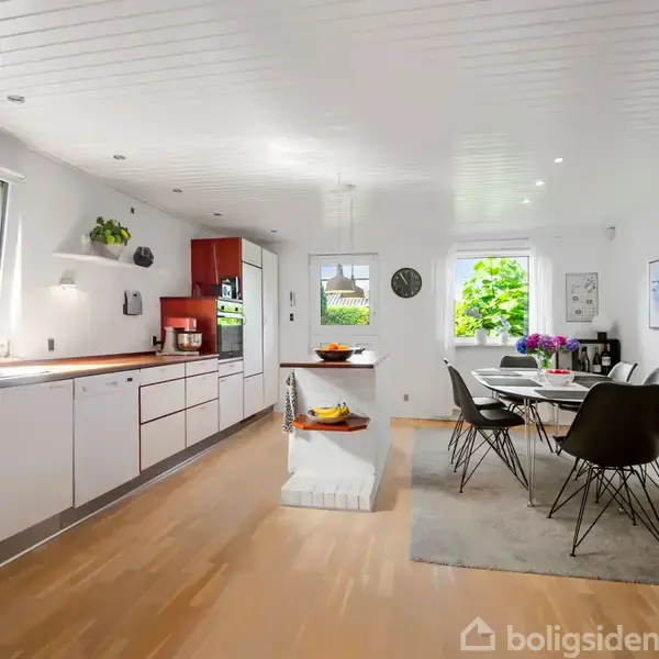 Et moderne køkken med lange bordplader og integrerede skabe, set med en spiseplads til seks personer. Store vinduer slipper lys ind, og der er kunst på væggene.