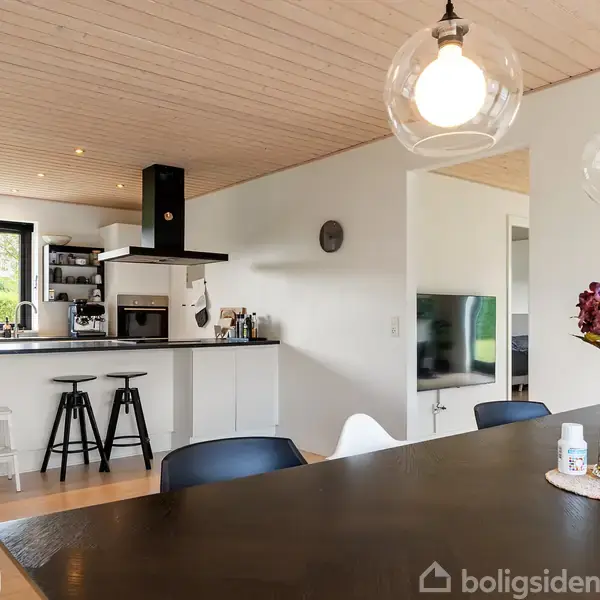 Et åbent køkken med hvide skabe har en køkkenø med sorte barstole. I nærheden er der et spisebord med en buket lilla blomster i en vase og moderne lamper hængende over.