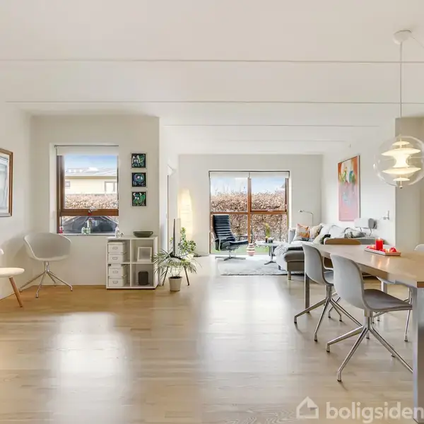 Stue med træbord og stole, lamper hængende over bordet, sofaområde i baggrunden, store vinduer og døre ud til terrasse, dekoreret med planter og kunstværker på væggene.