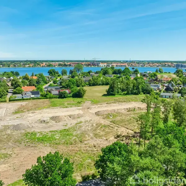 Åben mark med jordarbejde omringet af grønne træer og buske. I baggrunden ses en by med huse og bygninger ved en sø, under en klar blå himmel.