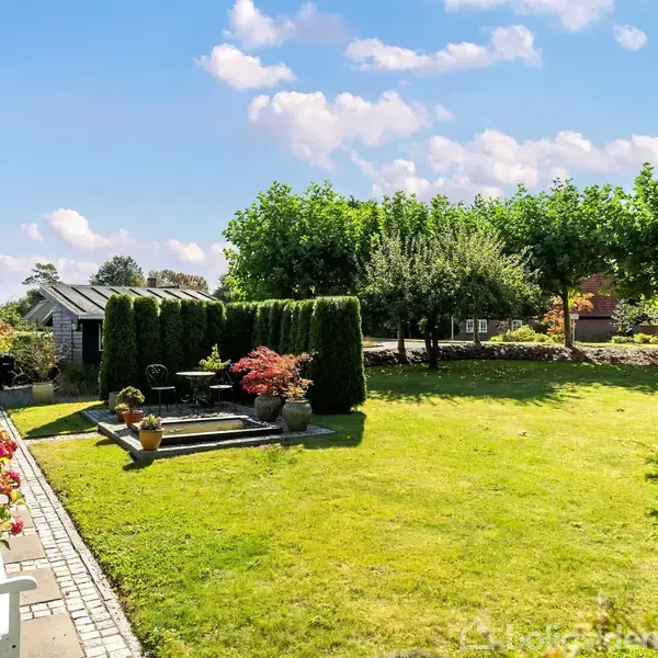 En have med græsplæne, blomsterbede og en stenbelagt terrasse med bord og stole, omgivet af frodige buske og træer. En hvid bænk står ved husmuren på venstre side.