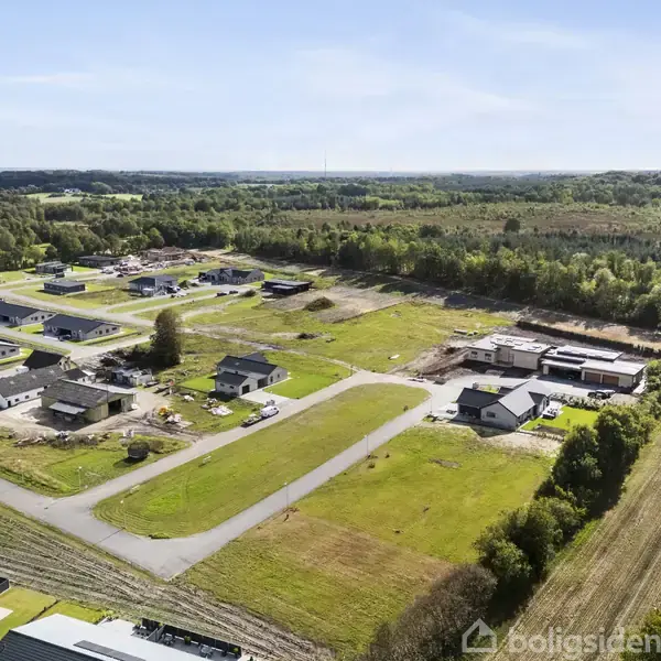 Luftfoto af et nybygget boligområde. Rækkehuse og villaer spredt omkring med grønne områder og skov i baggrunden. Stier og veje forbinder de forskellige sektioner.