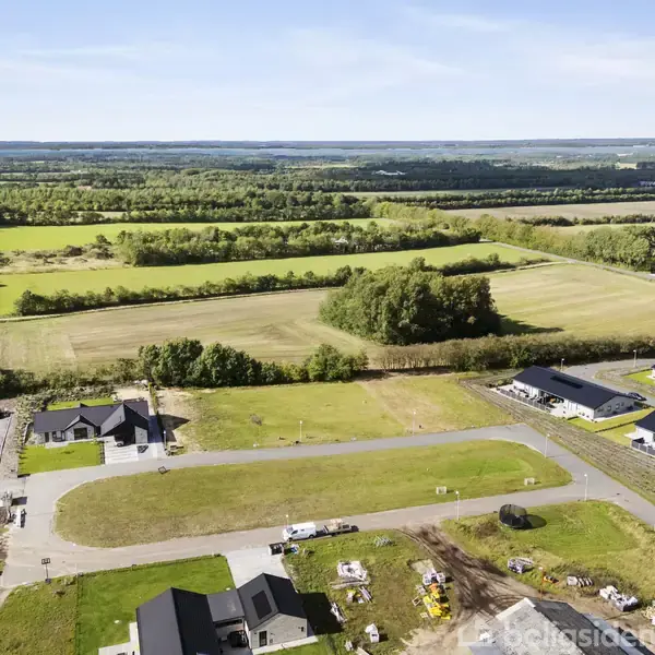 Luftfoto af et parcelhuskvarter under udvikling, hvor flere huse er under konstruktion. De omkringliggende omgivelser inkluderer åbne marker og træer, der strækker sig til horisonten.