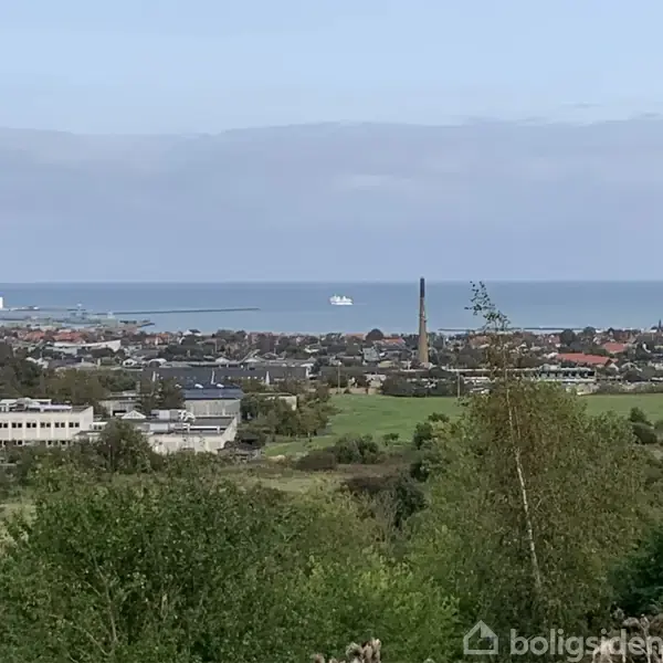 En by set fra en skovklædt bakketop, med huse, en høj skorsten, græsarealer og en hvid færge på vandet i horisonten under en blå himmel.