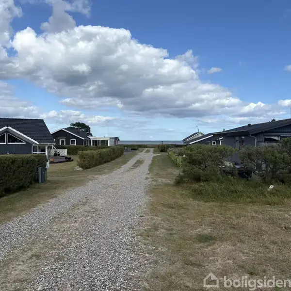 Grusvej strækker sig mellem to række af grå huse med sorte tage. Huse er omgivet af grønne hække under en blå himmel med hvide skyer.