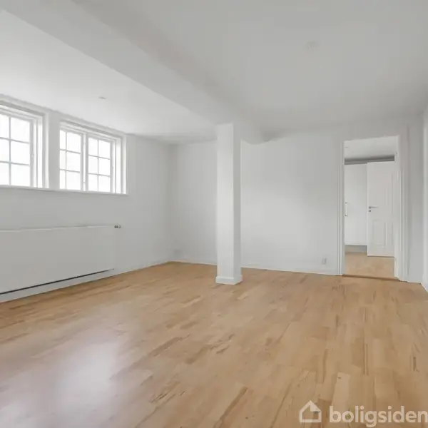 Et lyst rum med trægulv, hvor en radiator er placeret under vinduer med sprosser. Rummet er omgivet af hvide vægge og en enkelt søjle, og der er en døråbning i baggrunden.