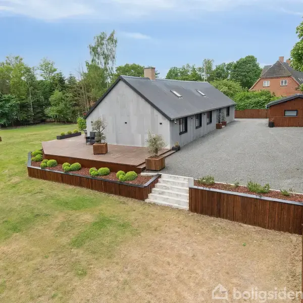 En grå bungalow med sort tag står på en stor grund. Bygningen har en træterrasse med planter omkring. Omgivet af en grusbelagt indkørsel og grønne træer.