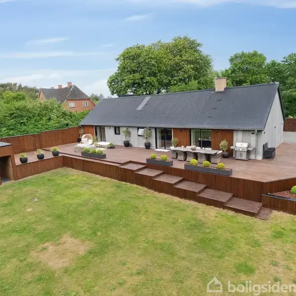 En moderne villa med fladt tag står på en stor træterrasse med havemøbler og grill. Haven er frodig med græsplæne og omkranses af grønne træer og buske.