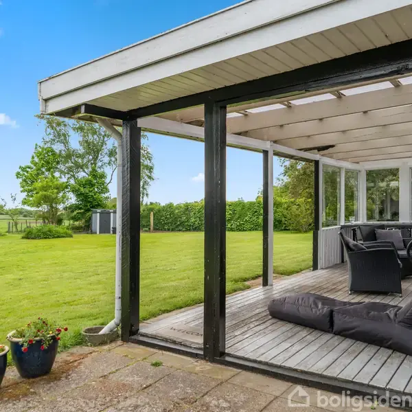 Overdækket terrasse med sorte stole og en liggepude, placeret på et trægulv. Omkring ligger en stor græsplæne omgivet af hække og træer under en klar blå himmel.