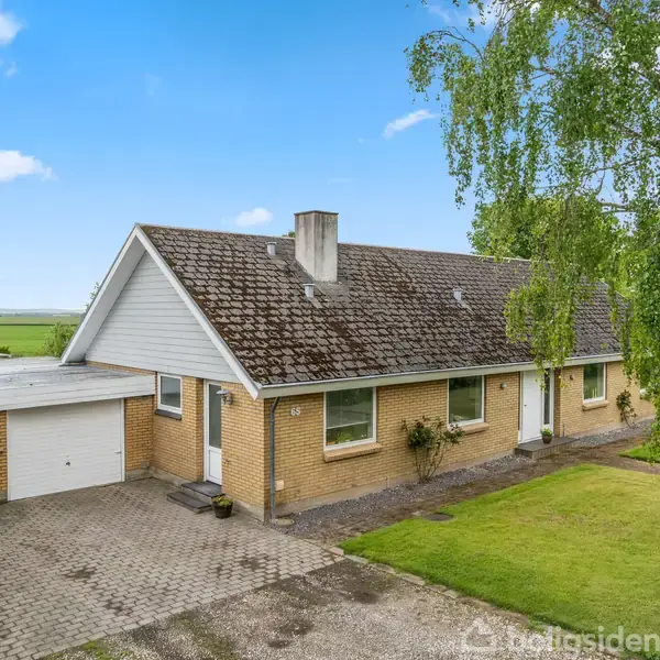 Huset er en gulstensbungalow med garage, stående i et landligt område med græsplæne og blomster. En indkørsel i belægning fører op til indgangen. Det er en solrig dag.