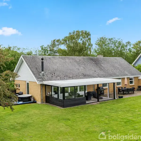 Et enkeltplanshus med gul murstensfacade og grått tag. Foran huset er en stor, grøn græsplæne omgivet af træer. Huset har en terrasse med glasoverdækning.