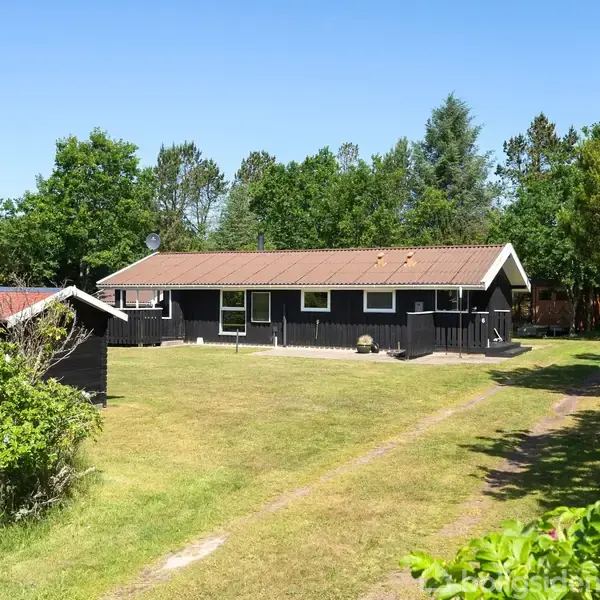 Et sort sommerhus med skodder står fredeligt på en græsplæne, omgivet af tætte, grønne træer under en klar blå himmel. En indkørsel fører til huset, som har en satellitantenne.