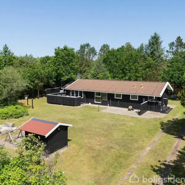 Et sort hus med veranda står omgivet af en stor græsplæne og tæt skov. Ved siden af findes et mindre udhus med rødt tag. Solskinsdag med blå himmel.