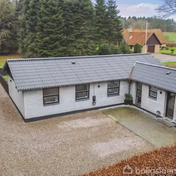 Et enkelt, hvidmalet hus med gråt tag står roligt på en grusindkørsel. Omkranset af høje, tætte træer, ligger det i et landligt område med grønne omgivelser og en nærliggende bygning.