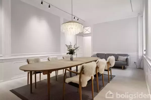 Langt spisebord med seks stole dækket af hvide skind omkring det. Fin lysekrone hænger over bordet. Bagved er en grå sofa i en rummelig stue med en lys, elegant indretning.