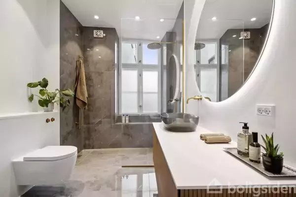 Et moderne badeværelse med en stor rund spejl og grå vask på træbord. Der hænger en plante over et toilet til venstre. Rummet har marmorfliser og stort vindue.