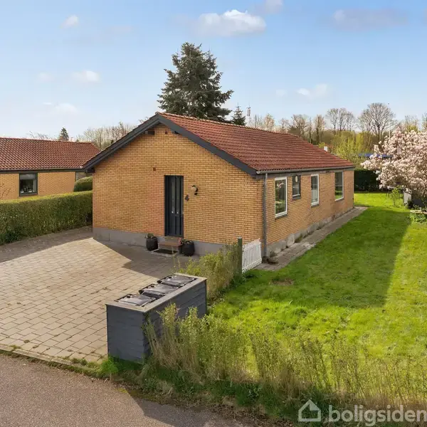 En gul murstensvilla med rødt tag står foran en flisebelagt indkørsel. Den er omgivet af en velplejet græsplæne, hække og et blomstrende træ i baggrunden under en blå himmel.