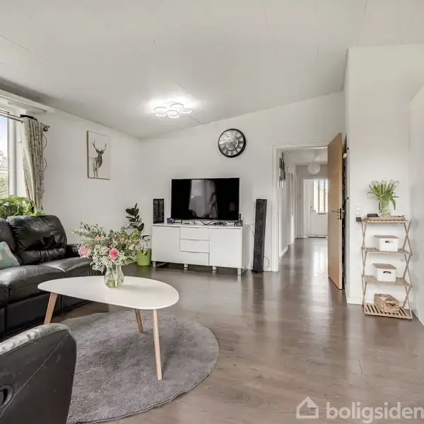 Sort sofagruppe står ved et rundt sofabord med blomster. Fjernsyn på et hvidt skab, væguret hænger over det. Lys stue med trægulv og stor vinduesudsigt.