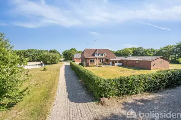 Rød murstensvilla med hvidt tag står midt i en grøn have, omgivet af grusvej og træer under en blå himmel.