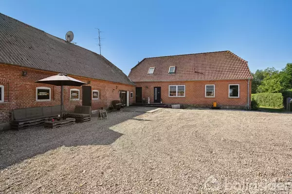 Murstensgård med to sammenhængende bygninger, står stille på en grusbelagt gårdsplads. Området er møbleret med et parasolcoveret siddehjørne. Baggrunden er et solrigt landskab.