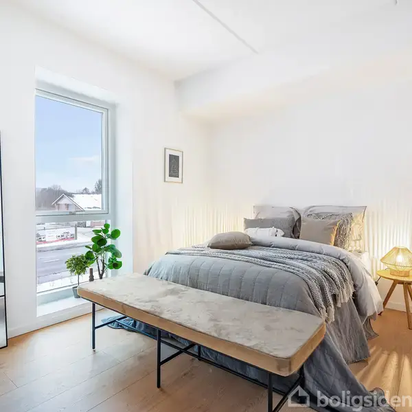 En dobbeltseng står i et lyst soveværelse med gråt sengetøj. Ved siden af er et lille natbord med en lampe. Et stort vindue giver udsigt til snedækkede omgivelser.