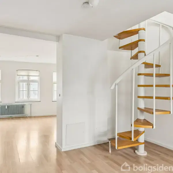 En hvid vindeltrappe fører opad i en lys stue med trægulv. Store vinduer med lys indstrømning skaber en rummelig atmosfære. Radiatorer ses i baggrunden ved vinduerne.
