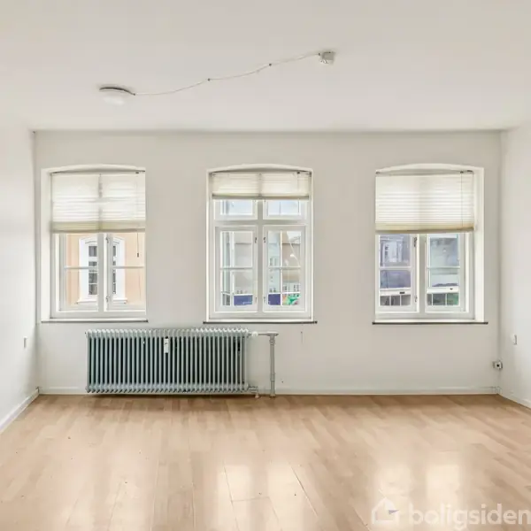 Et tomt rum med lyse trægulve har tre store vinduer med persienner på væggen. Under vinduerne står en radiator. Væggene er hvide, og der er ingen møbler.