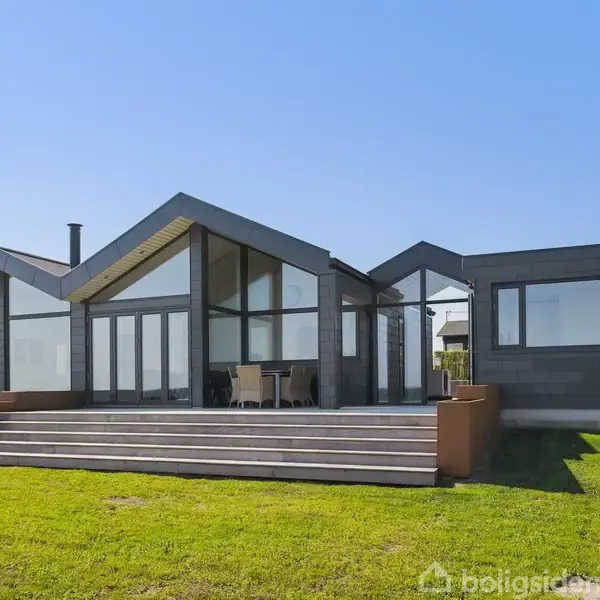 Et moderne, minimalistisk hus står med store glasfacader og en bred veranda, omgivet af en velplejet græsplæne under en klar blå himmel.