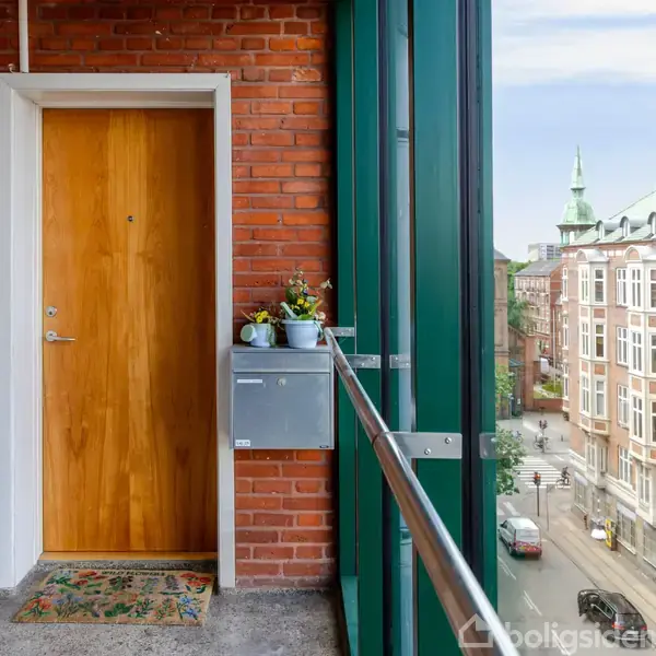 Dør i en rød murstensvæg, flankeret af en postkasse med blomster på. Foran løber en balkon med glasværn, der afslører en udsigt over en by med klassiske bygninger.