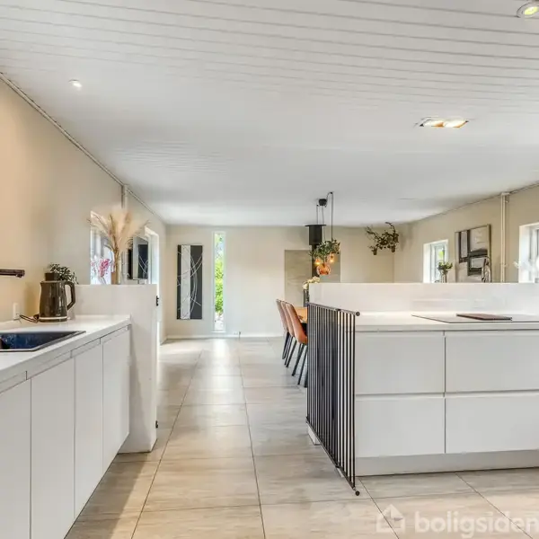 Moderne køkken med hvide skabe og køkkenø, fyldt med dagslys fra store vinduer og udsigt til grønne omgivelser udenfor. Rummets skandinaviske design skaber en lys og indbydende atmosfære.
