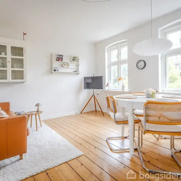 Stue med orange sofa står på hvidt tæppe. Rundt bord med stole ses tæt på to store vinduer. Vægskabe og hylder hænger på den hvide væg. En enkel vægklokke er også synlig.