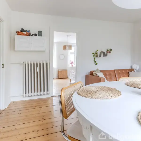 Rundt bord med dækkeservietter står i spisestue. I baggrunden ses en brun sofa, hvide vægge og træplankegulv. Rummet er lyst med minimalistisk indretning og grønne planter.