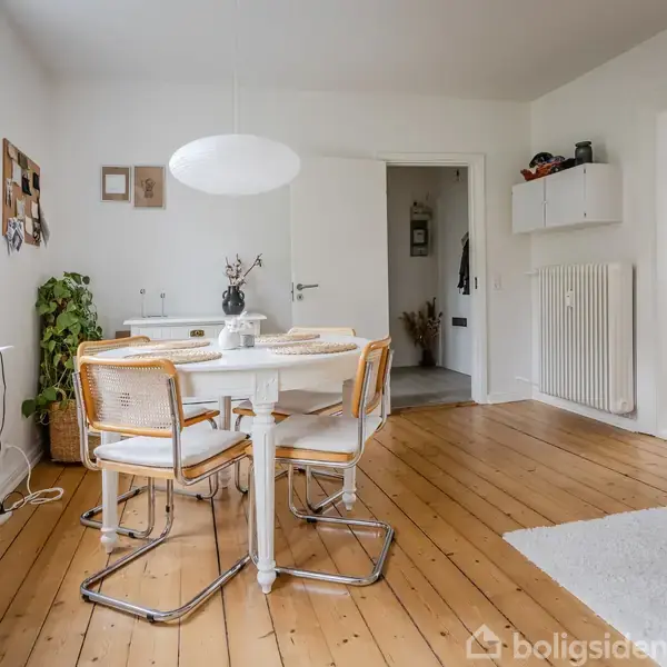 Et rundt spisebord med fire stole står i et lyst rum med trægulv. Der er hvide vægge, en lampe hænger over bordet, og en plante står i hjørnet.
