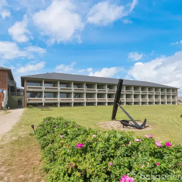 Anker står midt i en græsplæne med blomster omkring, foran en stor, grå bygning med altaner. Skyfri himmel og få skyer danner en lys, åben atmosfære.