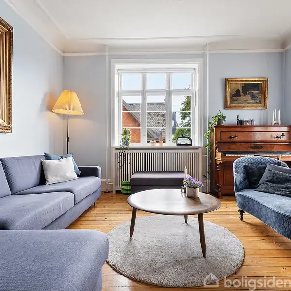 Stue med blå sofa og chaiselong ved et rundt træbord. Et klaver står ved væggen under et vindue. Omgivet af træmøbler, billeder, og en gulvlampe.