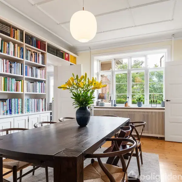 Et spisebord står i et lyst rum med en stor bogreol fyldt med bøger. Bordet har en vase med gule blomster. I baggrunden er der et vindue og en dør.