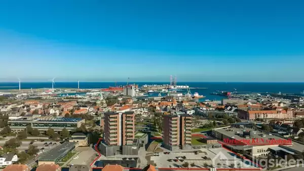 Høje bygninger står i forgrunden af en havneby, hvor adskillige vindmøller roterer i baggrunden ved kysten. Byens spredte huse og bygninger strækker sig ud mod havet.
