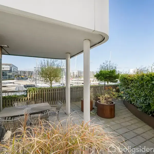 En møbleret terrasse med borde og stole omgivet af planter. Den er del af en bygning med udsigt over en marina og moderne lejlighedsbygninger i baggrunden.