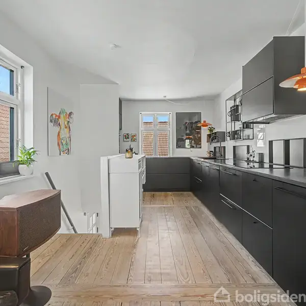 Sort køkken med moderne design, lang bordplade, integrerede apparater og træplankegulv. To vinduer for enden giver naturligt lys. Farverig kunst og planter tilføjer liv til rummet.