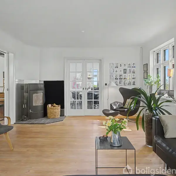 Et moderne stueinteriør med en sort lædersofa, en stol og et sofabord. Pejsen står til venstre. Et rum med glasdøre og mange billeder hænger på væggen ved siden af store vinduer.