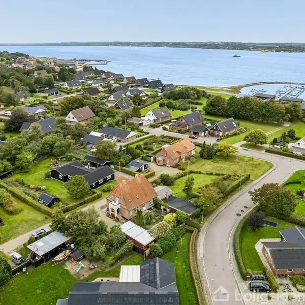 Flere huse ligger i et grønt, kuperet boligområde med veje, grønt græs og træer omkring. I baggrunden ses en stor sø eller fjord.