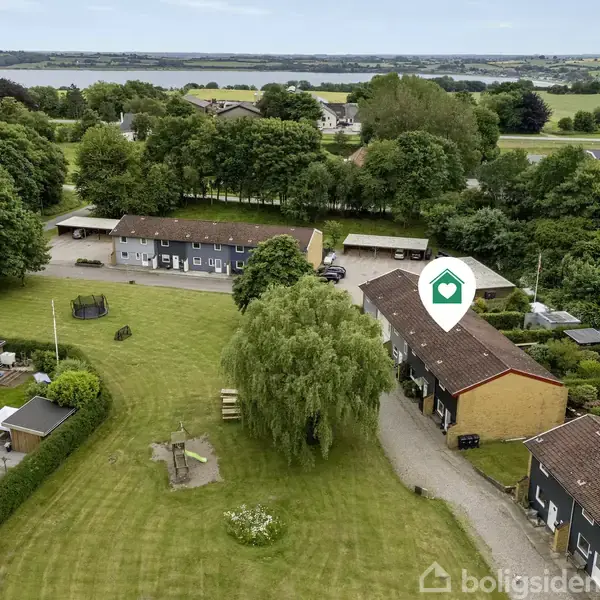 Rækkehuse ligger i en grøn, åben have, omgivet af træer. I baggrunden ses flere huse og en sø. Et lille ikon med et hus er placeret over det ene hus.