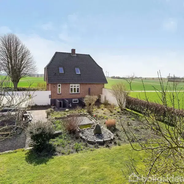 Rødt murstenshus med sort tag står i en have med græsplæne og buske. Landskabet omkring er åbent med marker og enkelte træer under en blå himmel.