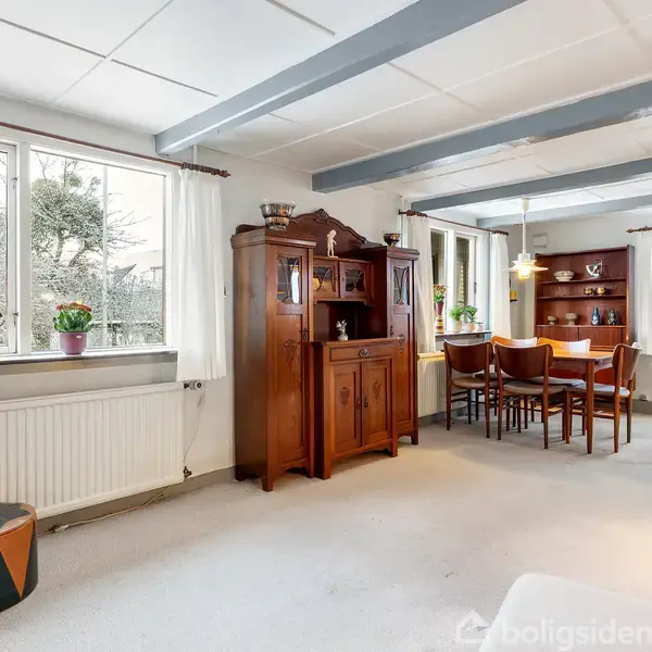 Et spisebord med stole står i et lyst rum med hvide vægge, træbjælker i loftet, og vinduer med gardiner. Der er en træskænk ved siden af bordet.